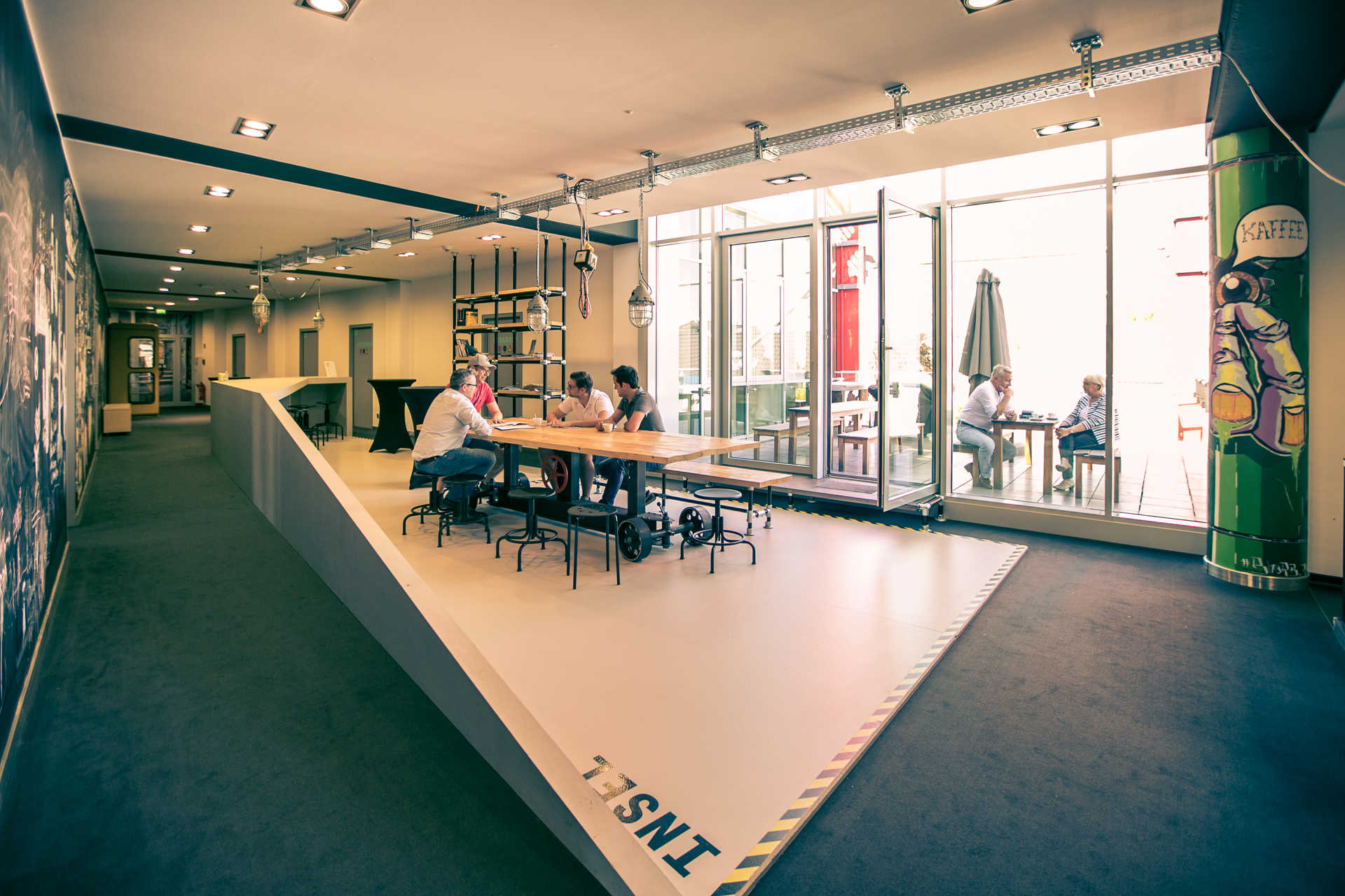 Mehere Personen sitzen an einem großen Essenstisch, der durch eine auffällige Kurbel hoch und runter gefahren werden kann.Im Hintergrund ein Bücherregal, sowie der Blick auf die Dachterrasse, wo sich 2 Personen an einem Tisch unterhalten.